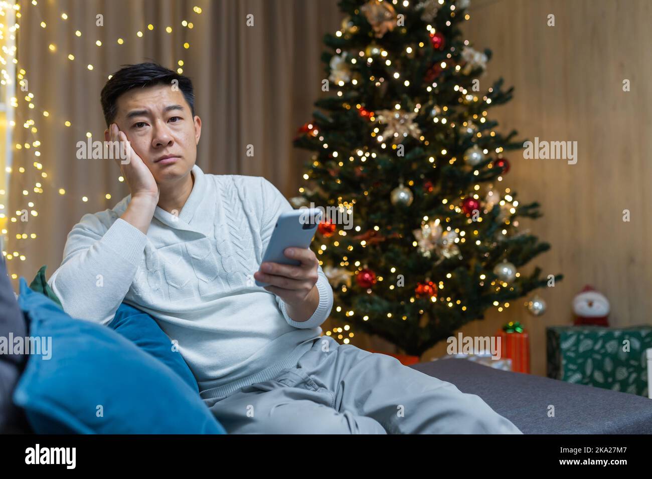 Trauriger und einsamer junger asiatischer Mann sitzt zu Hause auf dem Sofa in der Nähe des Weihnachtsbaums und hält das Telefon in den Händen. Warten auf einen Anruf, Benachrichtigung für den Urlaub. Stockfoto