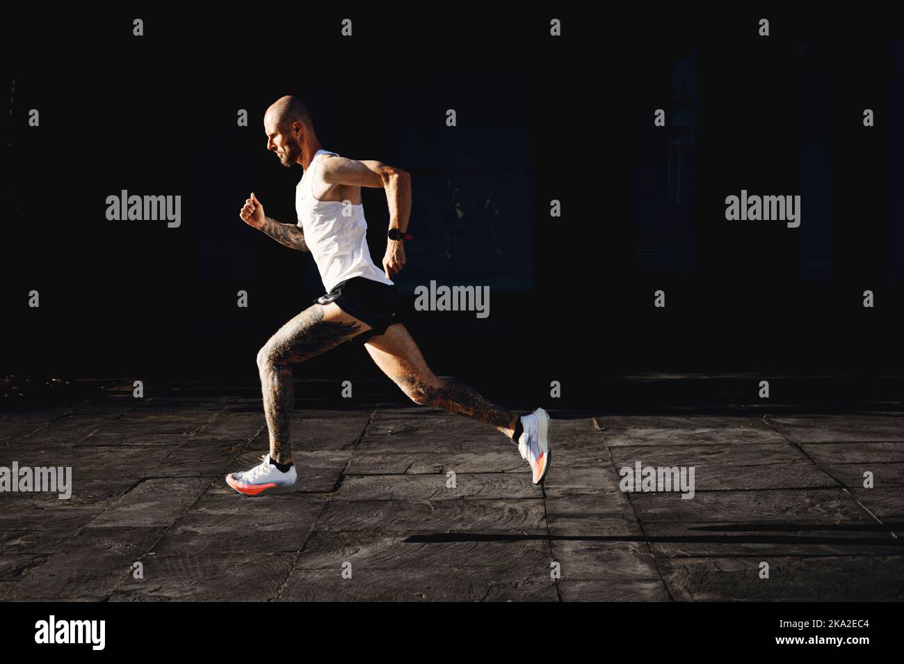 Kahlköpfiger junger athletischer tätowierte Mann läuft schnell im Freien, Sportler bereitet sich auf den Marathon vor, macht Cardio-Übungen im Freien, Seitenansicht Stockfoto