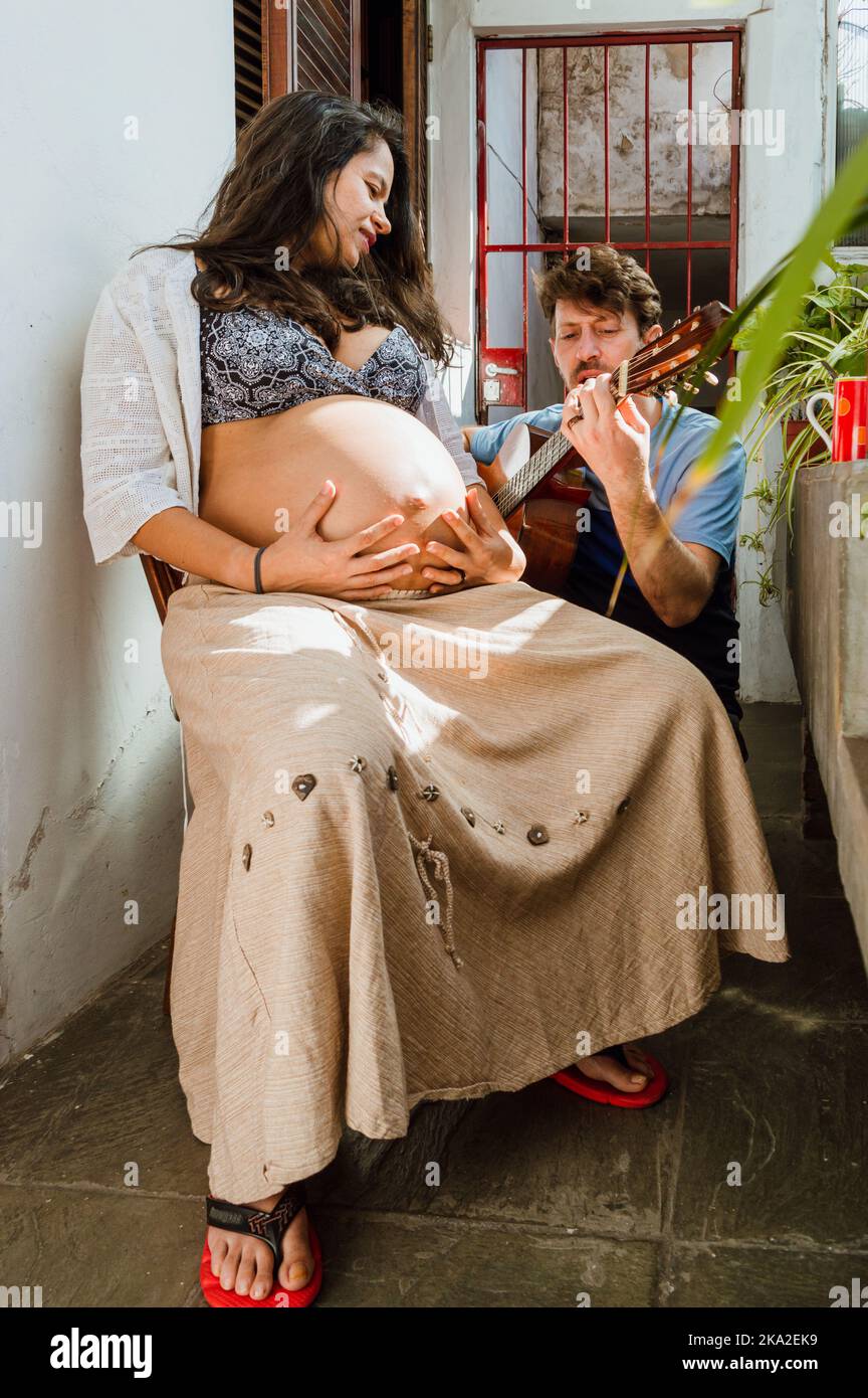 Ein junges heterosexuelles lateinisches Paar eines argentinischen Mannes mit Bart und kurzen Haaren und seiner schwanger brasilianischen Frau, das zu Hause Musik macht, Stockfoto