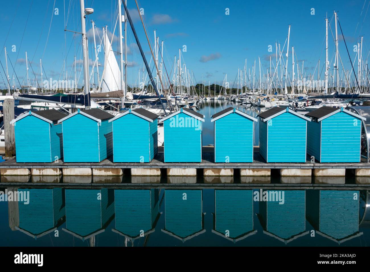 Blaue Hütten in Lymington Marina Hampshire England spiegeln sich im Meer mit Booten im Hintergrund Stockfoto