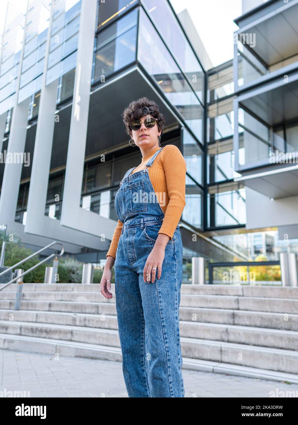 Von unten eine hispanische Dame in Sonnenbrillen und Jeans, die bei Tageslicht in der Nähe eines modernen Gebäudes auf die Kamera schaute Stockfoto