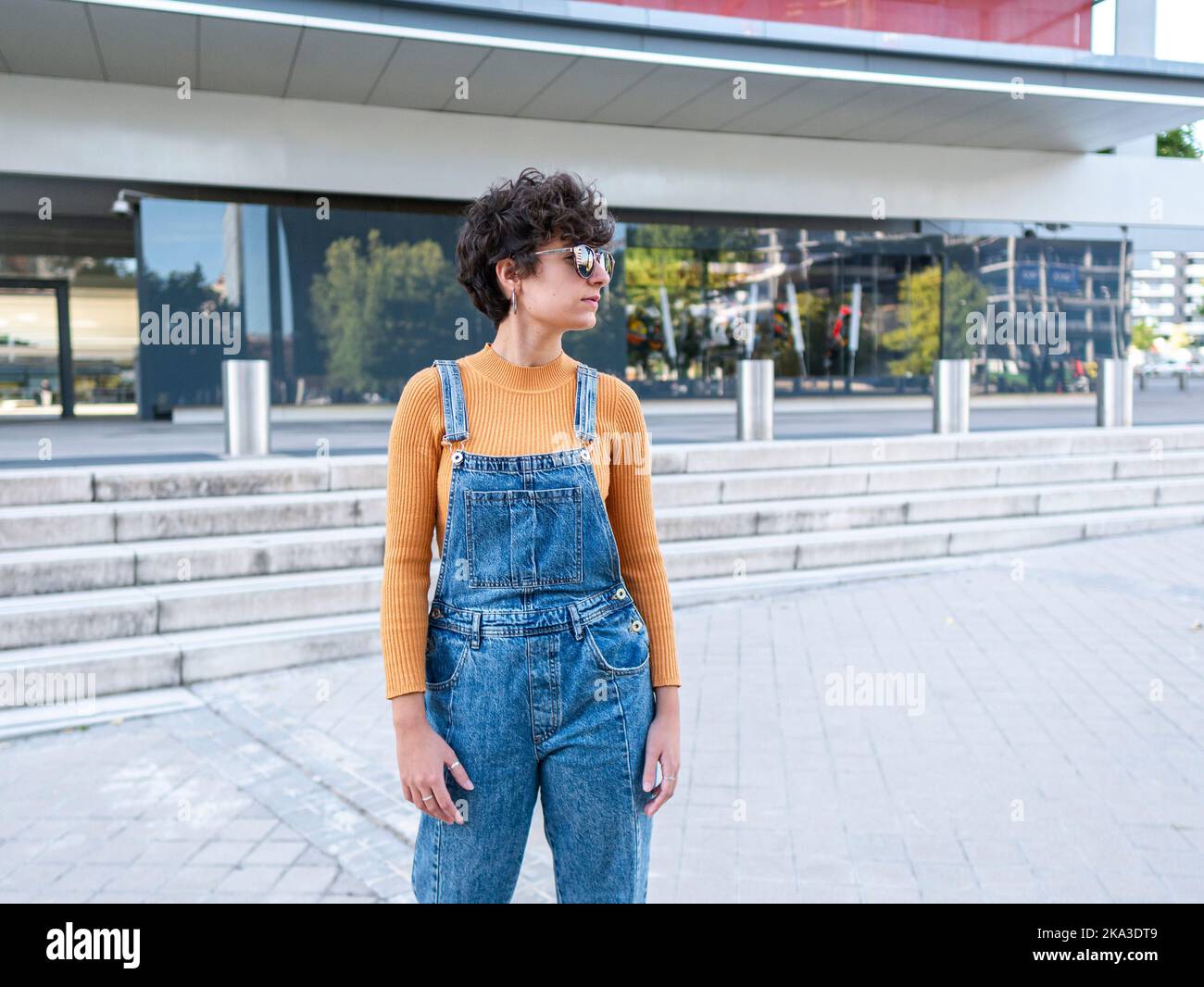 Die hispanische Dame in Sonnenbrillen und Jeans steht bei Tageslicht und blickt in die Nähe eines modernen Gebäudes Stockfoto
