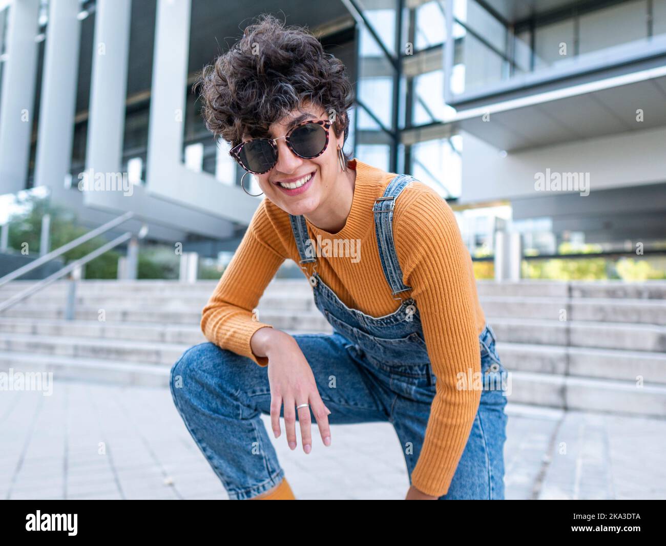 Lächelnde hispanische Dame in Sonnenbrillen und Jeans lehnte sich insgesamt auf den Arm und schaute bei Tageslicht in die Nähe eines modernen Gebäudes auf die Kamera Stockfoto