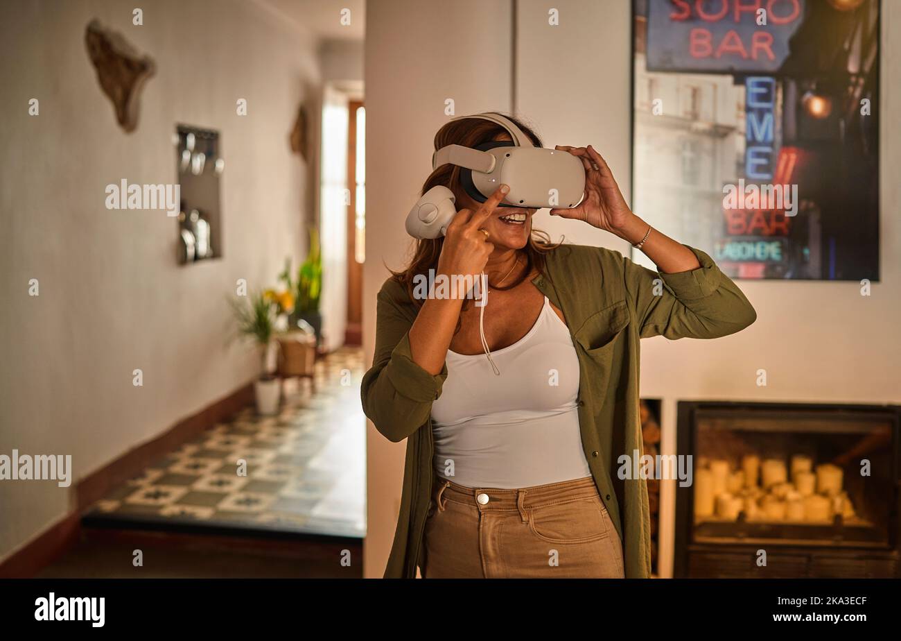 Fröhliche Frau mittleren Alters in stilvoller Kleidung lächelt und justiert VR-Helm, während Sie am Wochenende zu Hause den Cyberspace erkunden Stockfoto