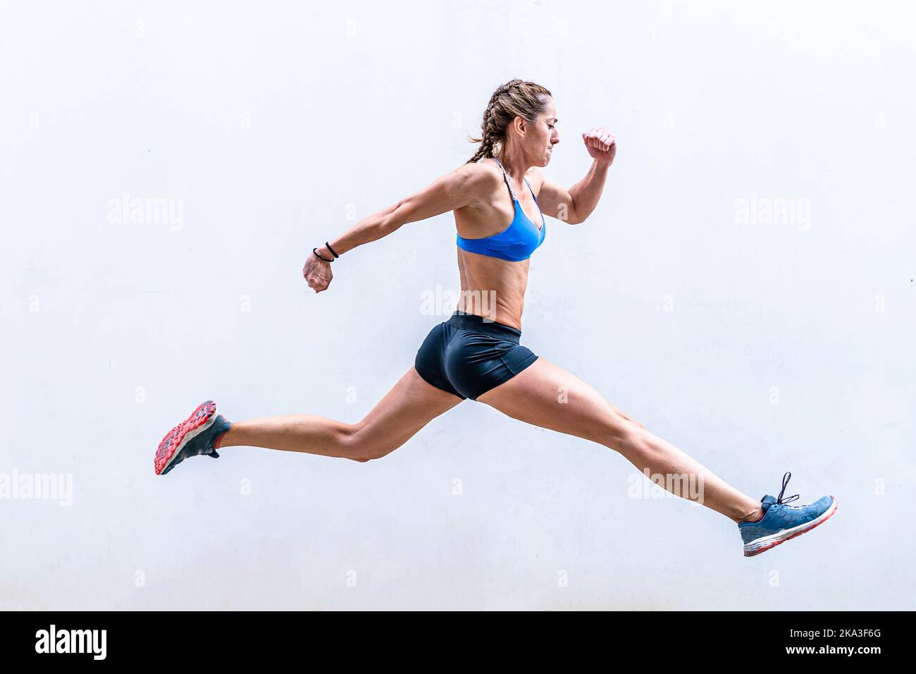 Ganzkörper bestimmt muskulösen weiblichen Athletinnen im aktiven Ohr heben Arm und springt gegen weiße Wand während intensives Fitness-Training Stockfoto