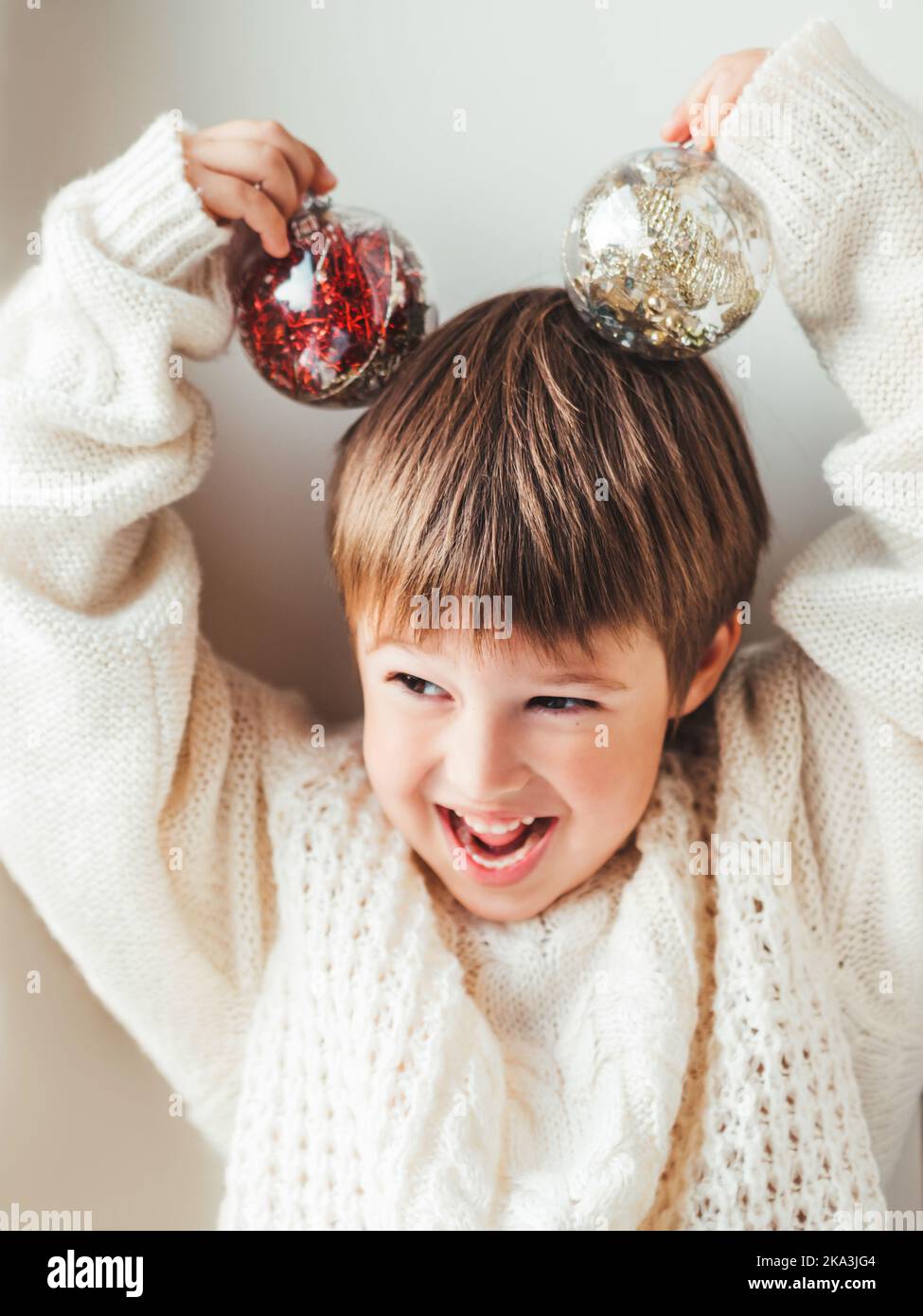 Kind mit dekorativen Kugeln für Weihnachtsbaum.Junge in Zopfmuster übergroße Pullover.gemütliches Outfit für kuscheliges Wetter.Transparente Kugeln mit roten, goldenen sp Stockfoto
