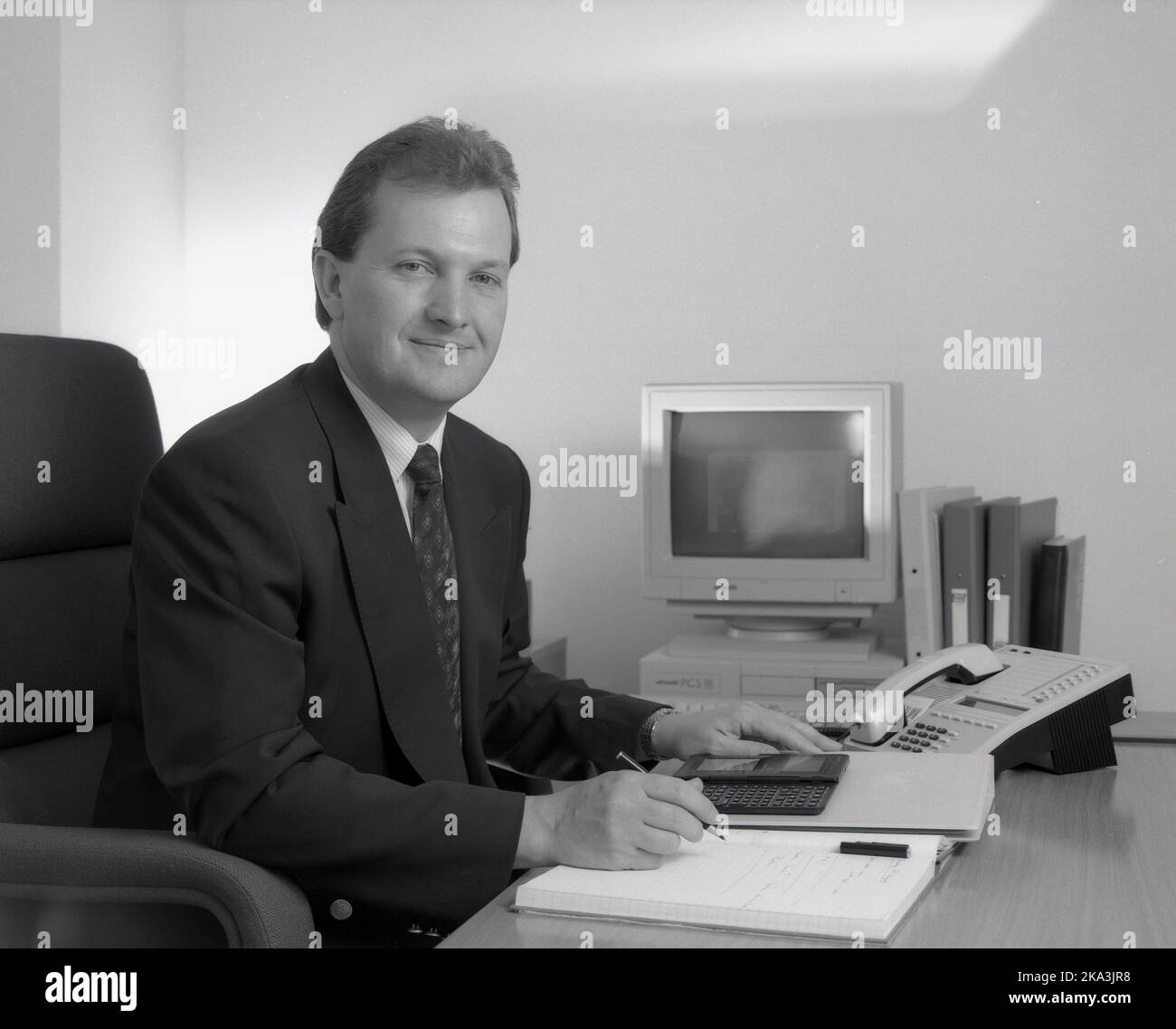 1991, historisch, ein geeigneter männlicher Geschäftsleiter, der mit einem Notebook, einem Faltblatt, einem Telefon und einem elektronischen Organisator - auch bekannt als persönlicher Digitaal-Assistent (PDA) - an seinem Schreibtisch sitzt. Ebenfalls auf dem Bild zu sehen, ein PC der damaligen Zeit, ein Olivetti PCS 286, mit VGA-Monitor und Tastatur. Dieser kleine Desktop-PC wurde um 1990 von Olivetti, einem führenden italienischen Computerhersteller, veröffentlicht und wurde sowohl für den privaten als auch den geschäftlichen Markt entwickelt. Er verfügt über einen R.A.M-Speicher von 1MB und ein 3,5-Zoll-Diskettenlaufwerk. Stockfoto