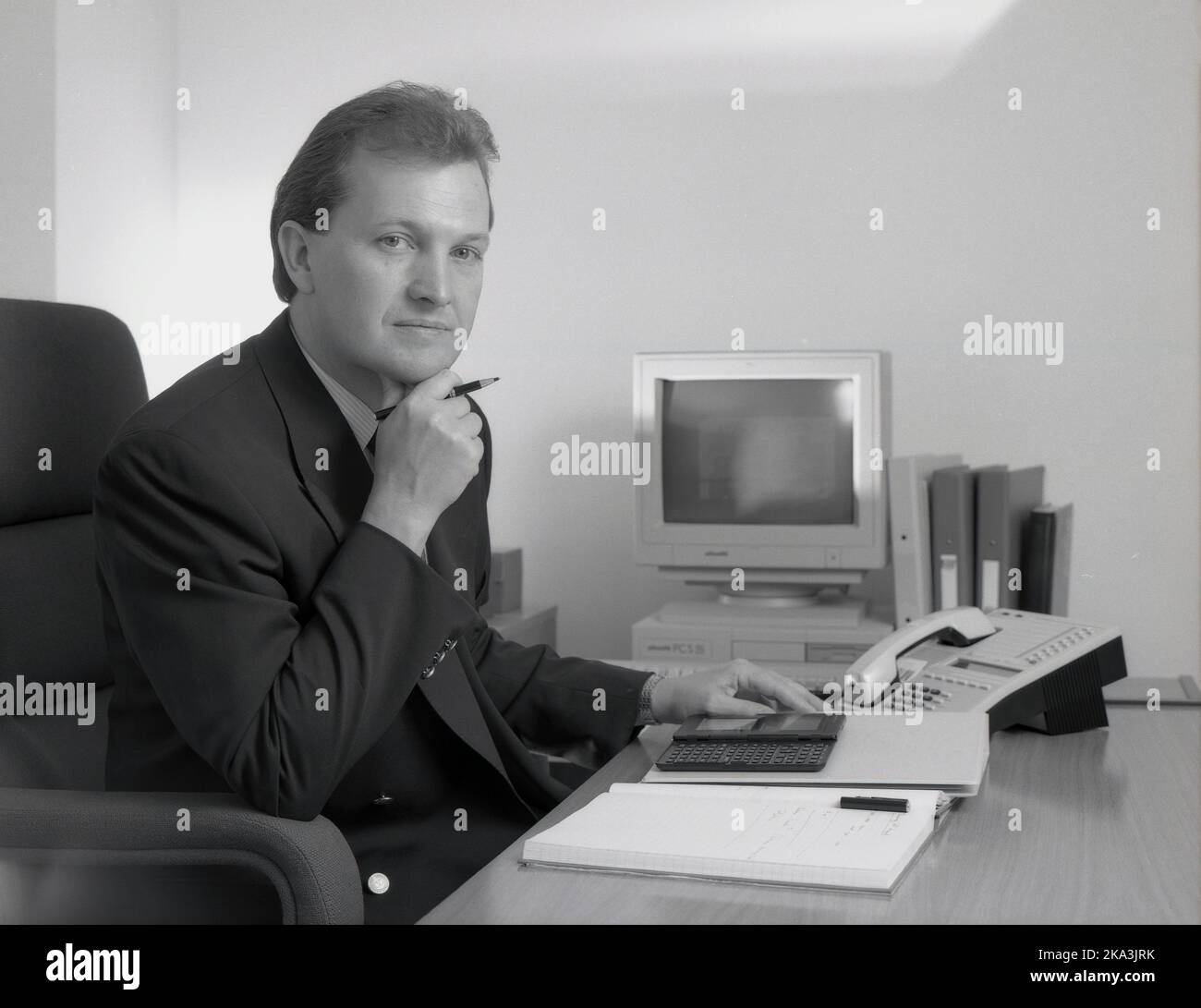 1991, historisch, ein geeigneter männlicher Geschäftsleiter, der mit einem Notebook, einem Faltblatt, einem Telefon und einem elektronischen Organisator - auch bekannt als persönlicher Digitaal-Assistent (PDA) - an seinem Schreibtisch sitzt. Ebenfalls auf dem Bild zu sehen, ein PC der damaligen Zeit, ein Olivetti PCS 286, mit VGA-Monitor und Tastatur. Dieser kleine Desktop-PC wurde um 1990 von Olivetti, einem führenden italienischen Computerhersteller, veröffentlicht und wurde sowohl für den privaten als auch den geschäftlichen Markt entwickelt. Er verfügt über einen R.A.M-Speicher von 1MB und ein 3,5-Zoll-Diskettenlaufwerk. Stockfoto