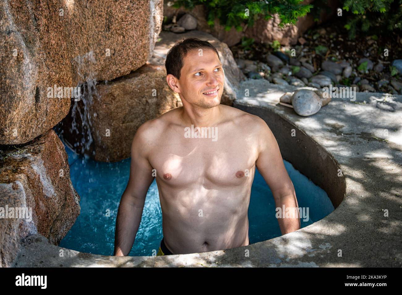 Junger, fröhlich lächelnder Mann schwimmt im japanischen Spa-Wasserfall-Steinpool mit farbenfroh blauem Wasser in den heißen Quellen Japans Onsen Stockfoto