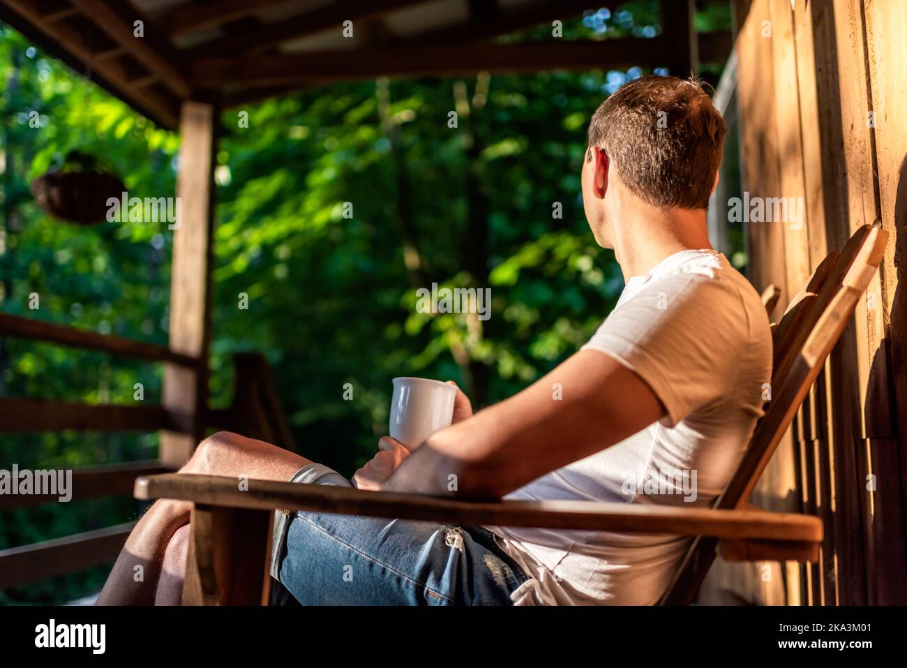 Mann, der auf einem Schaukelstuhl auf der Veranda des rustikalen Landhaushauses aus Holzhütte sitzt, den Sonnenaufgang am Morgen beim Kaffee aus der Tasse zu beobachten Stockfoto