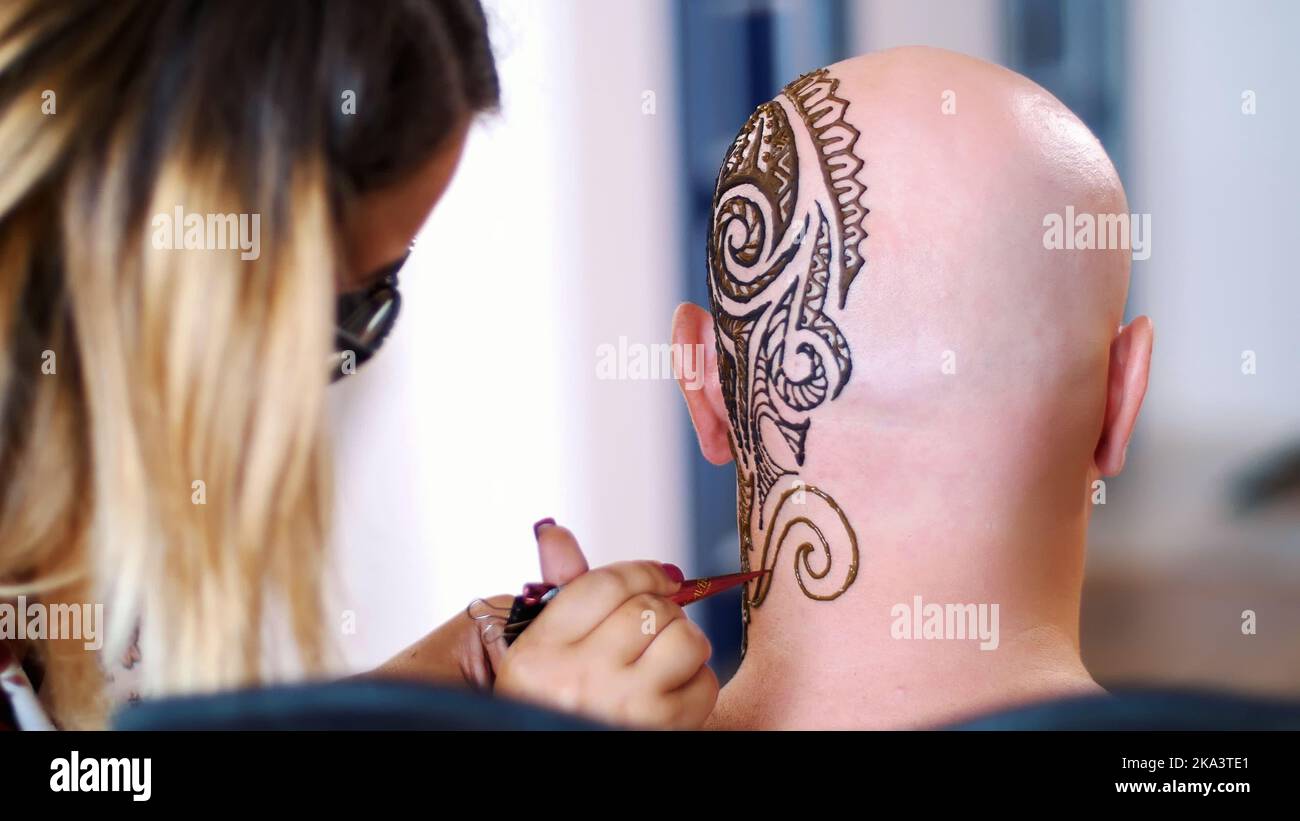 Ein Mädchen, Tattoo-Meister, mehendi Künstler macht Zeichnung von Henna-Tattoo auf der Haut von kahlem kaukasischen Mann, Schulter, Hals.die Lösung von Henna trocknet auf der Haut. Hochwertige Fotos Stockfoto