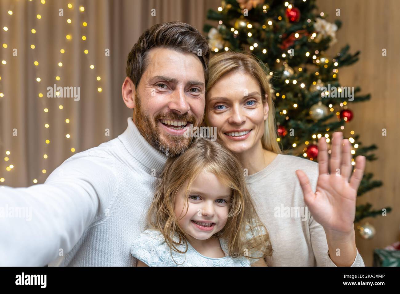 Weihnachtsfamilie, die mit einem Telefonvideoanruf telefoniert, Ehemann, Ehefrau und Tochter, die auf die Smartphone-Kamera schauen und am Telefon sprechen, um Freunde zu Weihnachten und Neujahr zu Hause in der Nähe des Weihnachtsbaums zu begrüßen. Stockfoto