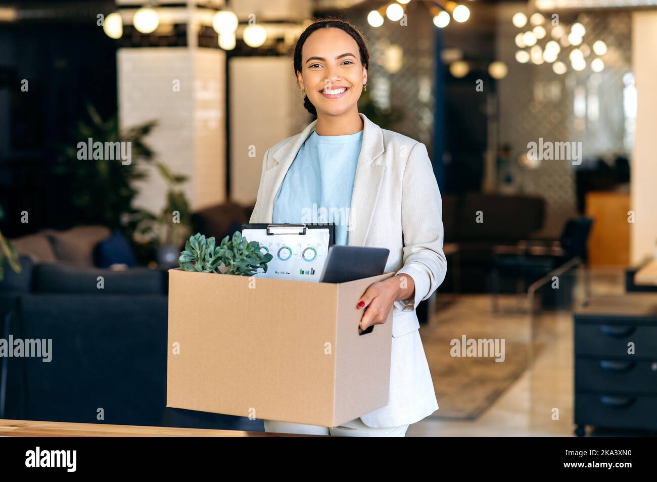 Neuer Job. Eine begeisterte, positive Mischlingsfrau, Auszubildende, die einen Karton hält, im modernen Büro steht und lächelnd die Kamera anschaut, hat ihren ersten Arbeitstag in einem neuen Arbeitsplatz Stockfoto