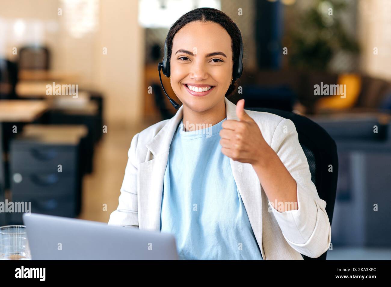 Wunderschöne, positive, selbstbewusste brasilianerin oder Latino-Frau mit Headset, Callcenter-Agent, Support-Servicemitarbeiter, sitzt im Büro, blickt auf die Kamera, zeigt eine Daumen-hoch-Geste und lächelt freundlich Stockfoto