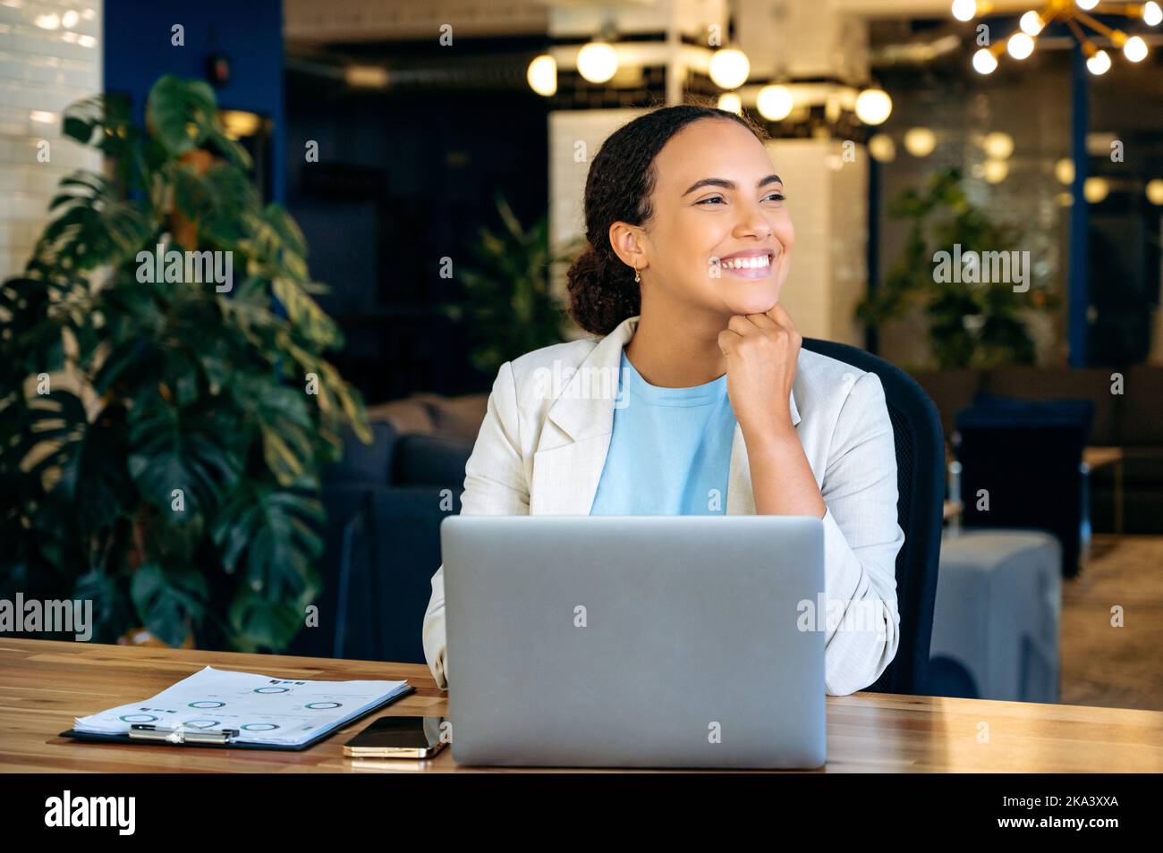 Glückliche erfolgreiche Mixed-Race-Geschäftsfrau, Programmiererin, Rekrutierung, in einer stilvollen Business-Kleidung, sitzt am Schreibtisch im Büro vor einem Laptop, wegschauen, denken, träumen, lächeln Stockfoto