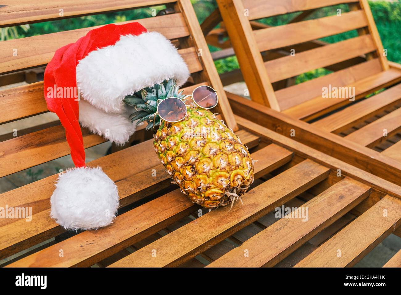Seitenansicht der entspannenden lustigen Ananas mit Sonnenbrille und Weihnachtsmannhut liegt bei sonnigem Wetter in den Tropen auf einer hölzernen Sonnenliege. Weihnachten in den Tropen Stockfoto