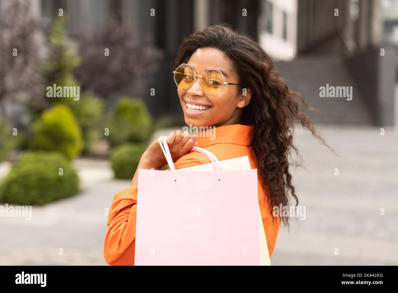 Porträt einer lächelnden jungen afroamerikanischen Frau in Casual und Brille mit Tasche mit Käufen, Blick auf die Kamera Stockfoto