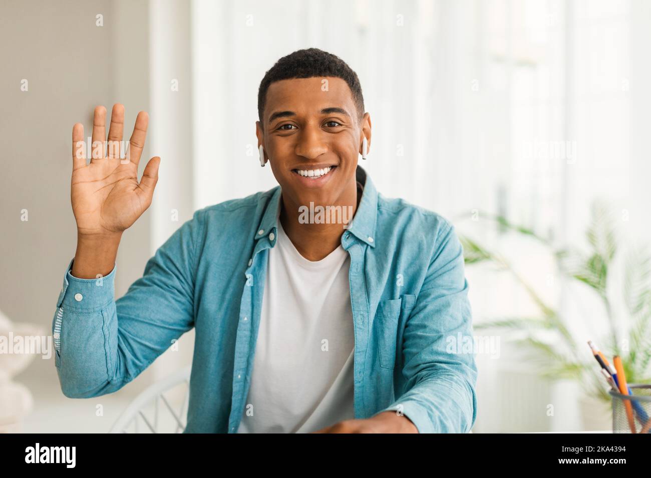 Fröhlicher junger afroamerikanischer Mann in zwangloser Atmosphäre mit kabellosen Kopfhörern, die die Hand schwenken und die Kamera betrachten Stockfoto
