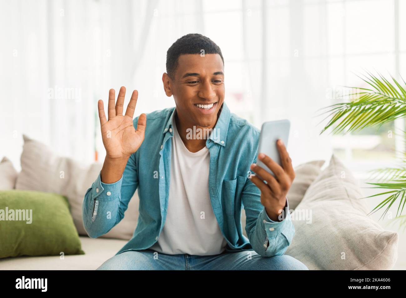Ein schöner junger afroamerikanischer Mann, der mit der Hand winkt und im hellen Wohnzimmer auf das Smartphone schaut Stockfoto