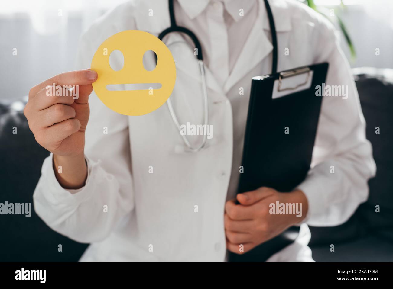 Nahaufnahme einer Ärztin in weißer Uniform mit Stethoskop, das ein gelbes trauriges Gesicht mit Papier hält, und einem schlechten Smiley, der auf dem Sofa sitzt. Feedback-Bewertung und Kundenrev Stockfoto