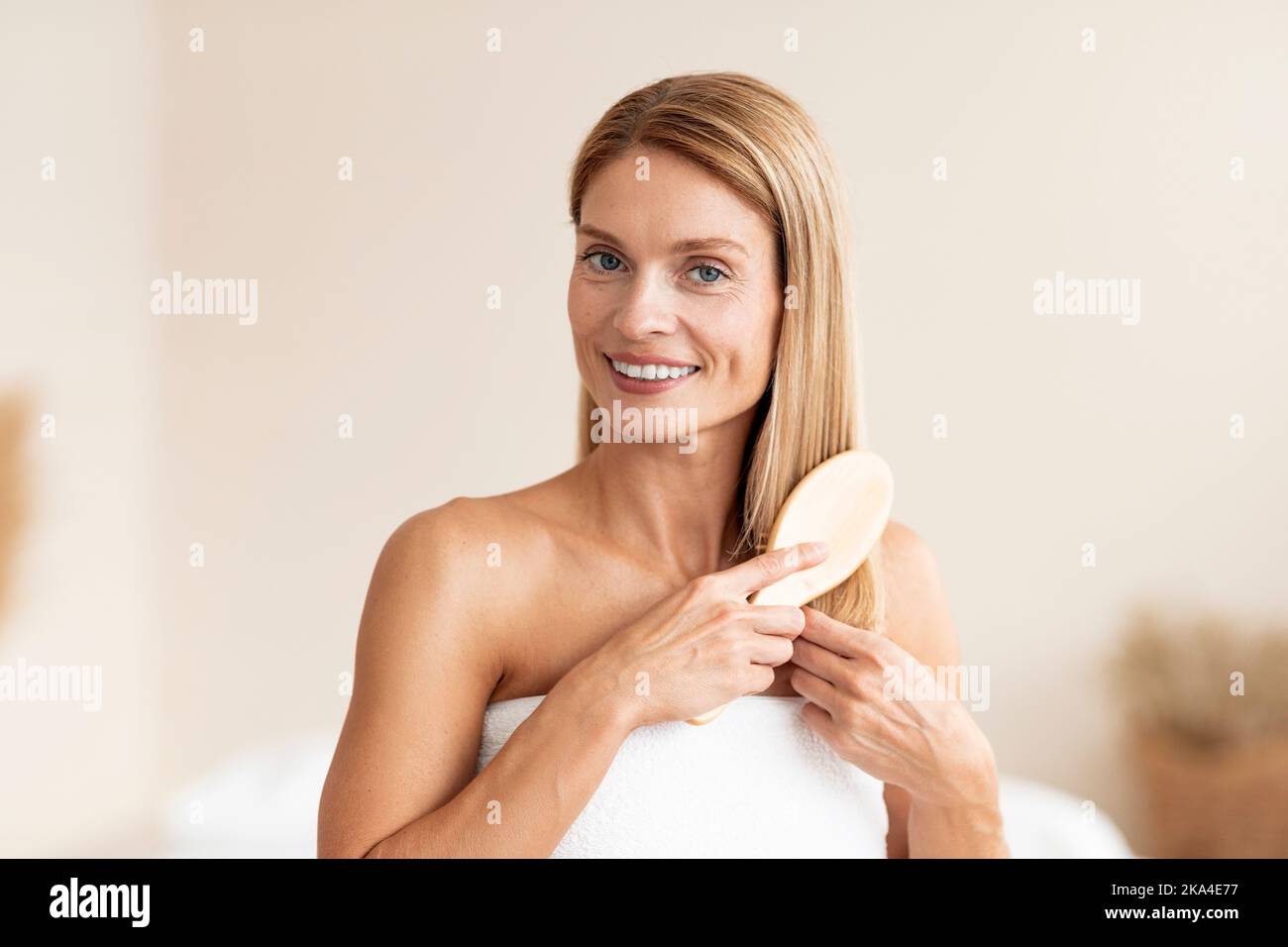 Morgendliche Schönheitsroutine. Attraktive reife Frau, die ihr blondes Haar mit Kamm putzt und in ein Badetuch gehüllt steht Stockfoto