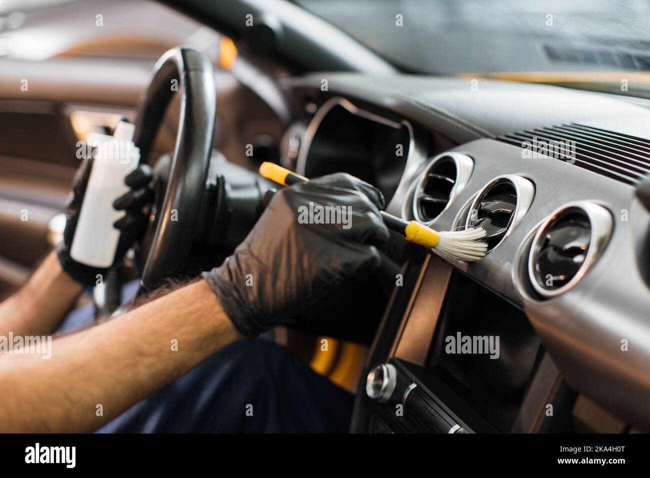 Beschnittenes Bild von Händen eines jungen männlichen Arbeiters in schwarzen Schutzhandschuhen, Reinigung des Armaturenbretts mit weicher Bürste und spezieller antistatischer Kunststoff-Ziermilch. Stockfoto