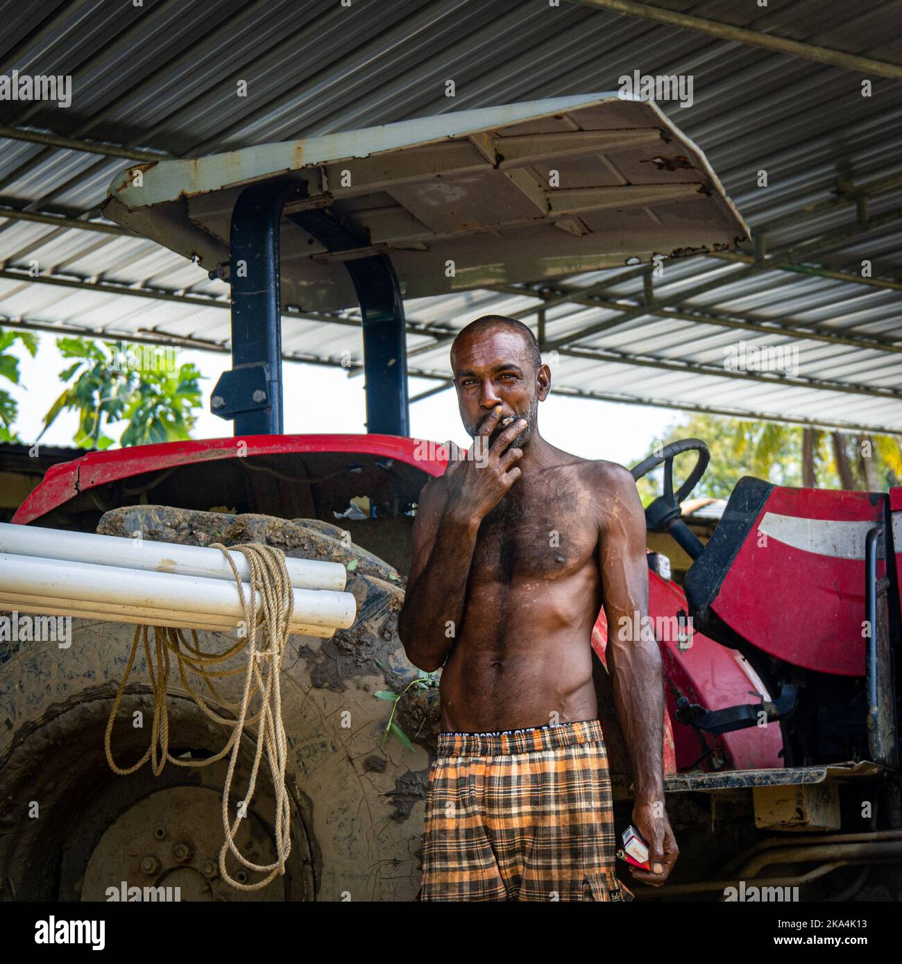 Ein schwarzer Mann, der vor seinem roten Traktor steht und eine Zigarette raucht Stockfoto