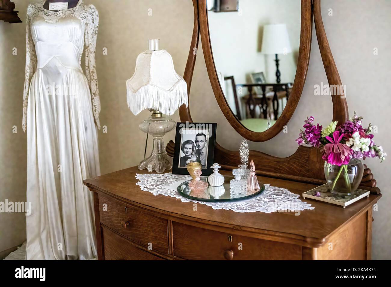 Antike Kommode und Spiegel mit einer Auswahl an Parfümflaschen, Hochzeitsfotos, Blumen, Lampe und einem Hochzeitskleid in der Ecke im Brautzimmer. Stockfoto