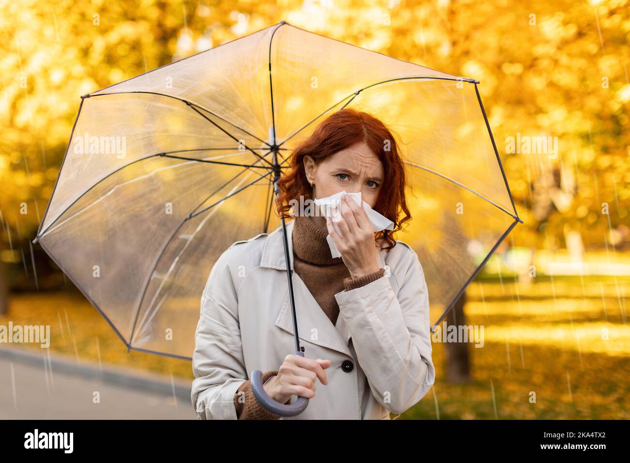 Traurige seriöse junge europäische rothaarige Dame im Regenmantel mit Regenschirm bläst ihre Nase auf Serviette, läuft im Regen Stockfoto