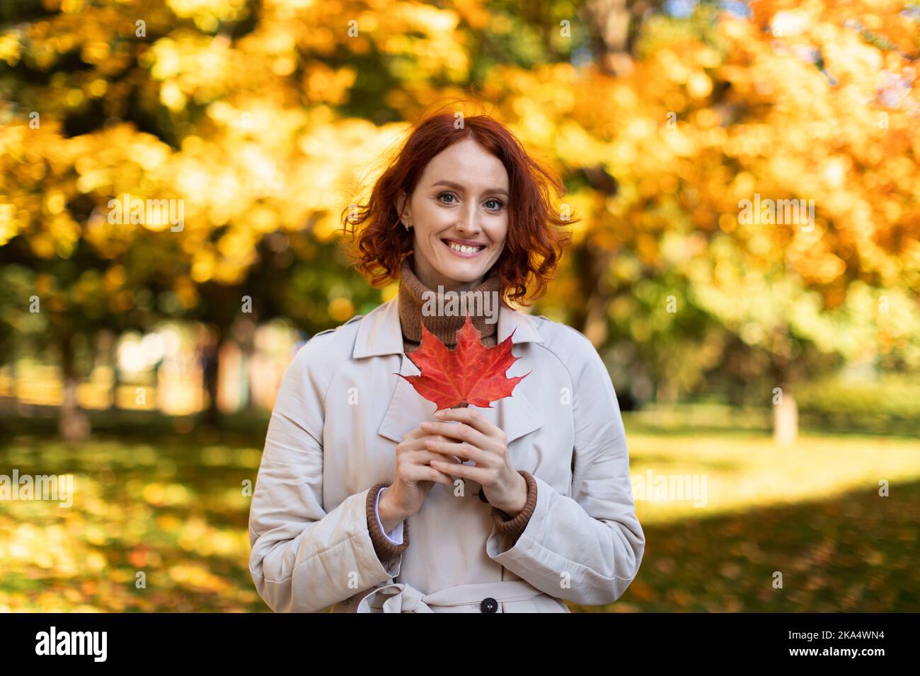 Porträt einer lächelnden, hübschen jungen europäischen rothaarigen Dame im Regenmantel halten im Herbst im Stadtpark ein rotes Blatt Stockfoto