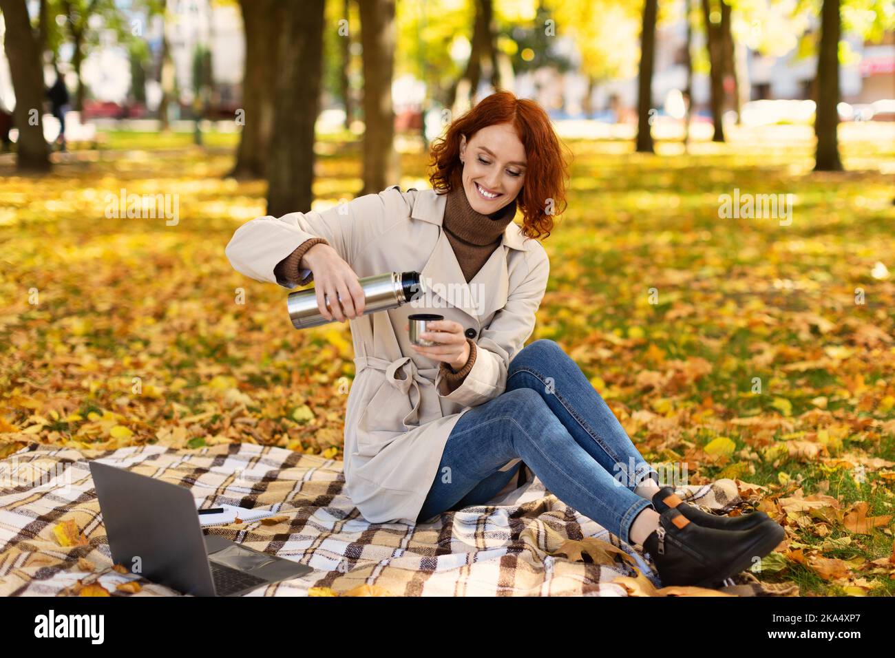 Fröhliche, hübsche junge, rothaarige Frau in Regenmantel mit Computer gießt Getränk aus Thermoskanne Stockfoto