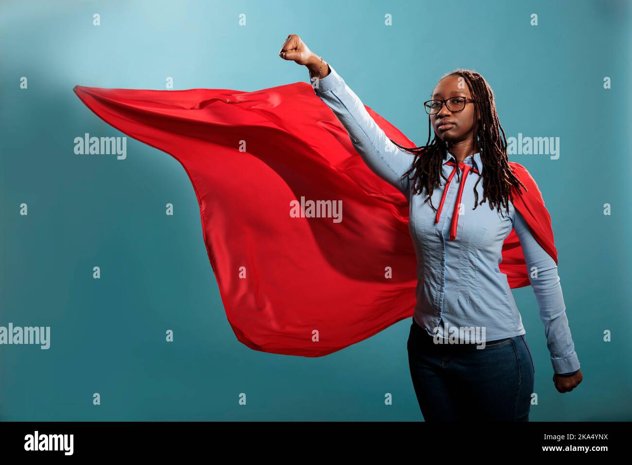 Starker Justizverteidiger mit Superkräften, die die Kamera mit Faust in der Luft betrachten. Tapfere und stolz aussehende junge Erwachsene Superheldin, die sich wie ein fliegender Held verhält, während sie einen mächtigen Umhang auf blauem Hintergrund trägt. Stockfoto