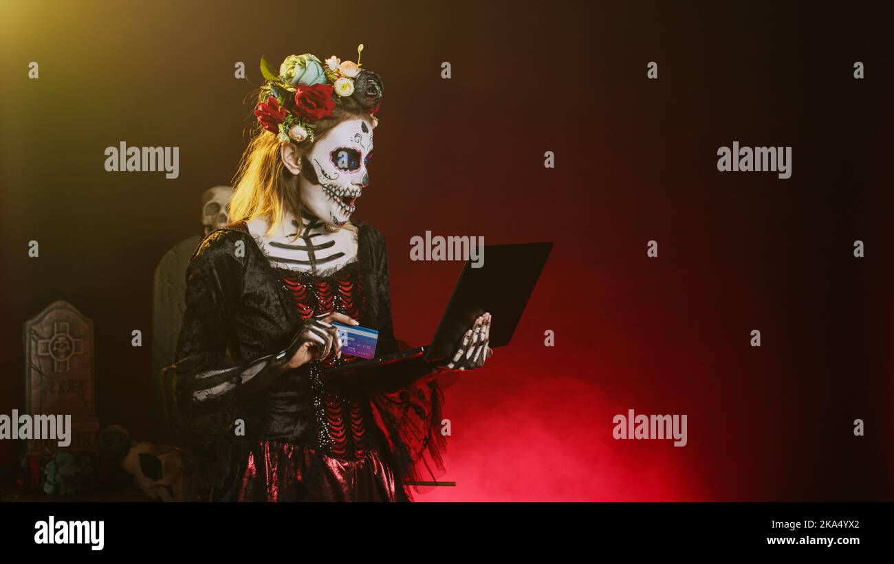 Halloween-Modell Online-Shopping mit Kreditkarte, mit Laptop, um elektronische Zahlung zu machen. Sieht aus wie die Dame des heiligen Todes oder santa muerte, die Internettransaktion auf dem pc bezahlt. Stockfoto