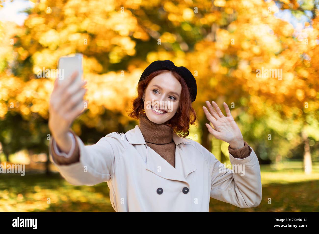 Lächelnde junge europäische rothaarige Dame in Regenmantel und Baskenmütze macht Selfie oder Video auf dem Smartphone, winkende Hand Stockfoto