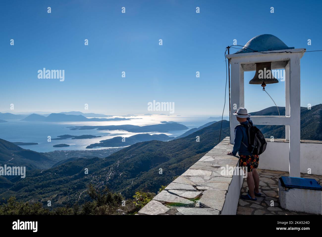 Ein Tourist an einem Glockenturm, der den Blick von der Bergspitze auf das Meer und die Inseln darunter genießt Stockfoto