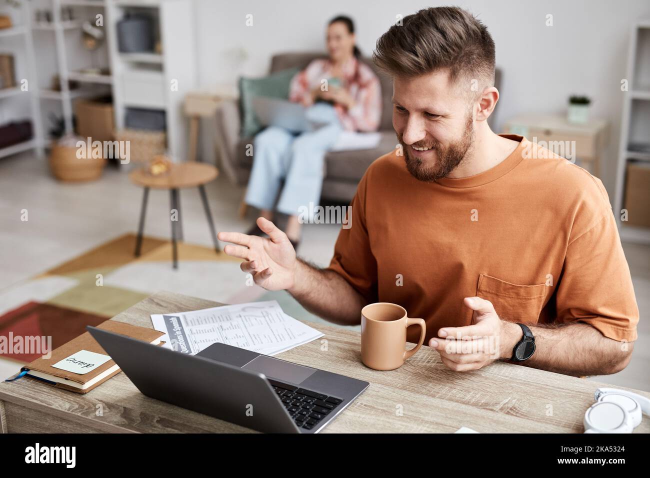 Junger lächelnder Mann in Casualwear, der zu Hause vor dem Laptop sitzt und während des Online-Unterrichts mit seinem Tutor kommuniziert Stockfoto