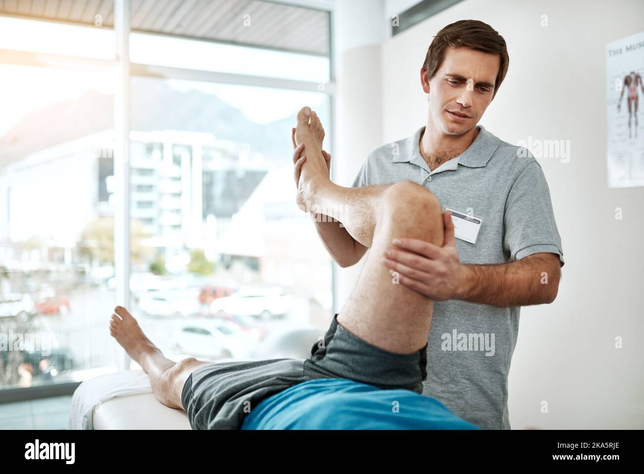 Tiefe Atemzüge. Ein junger männlicher Physiotherapeut hilft einem Klienten mit Beinübungen, der auf einem Bett liegt. Stockfoto