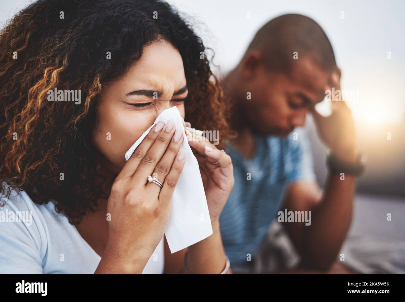 Sie kann einfach nicht aufhören zu niesen. Eine junge Frau bläst ihre Nase mit ihrem Freund, der im Hintergrund geniert aussieht. Stockfoto