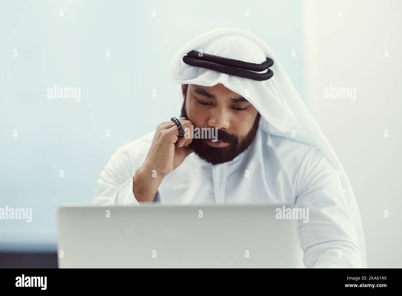In schwierigen Zeiten lehnt er sich an seinen Glauben. Ein junger Geschäftsmann, der in traditioneller islamischer Kleidung gekleidet ist und Gebetsperlen hält, während er in seinem Büro arbeitet. Stockfoto