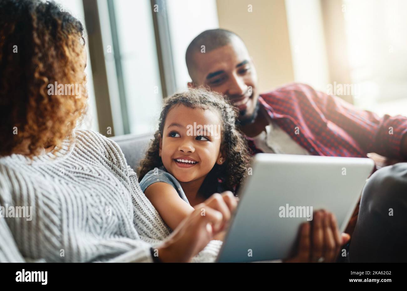 Darf ich das drücken, Mama? Ein entzückendes kleines Mädchen und ihre Eltern benutzen ein digitales Tablet zusammen auf dem Sofa zu Hause. Stockfoto