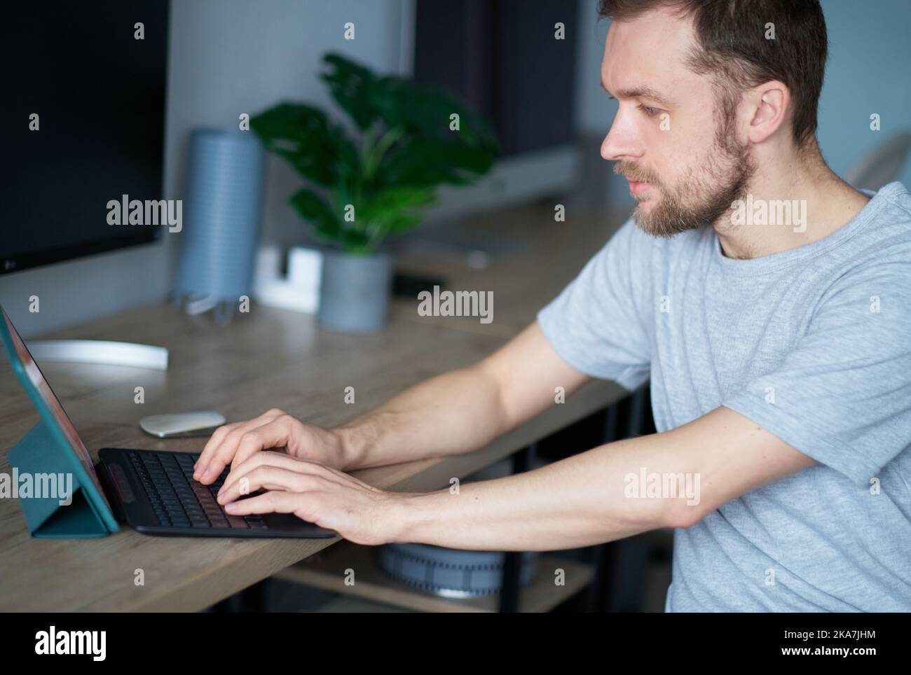 Fröhlicher bärtiger männlicher Software-Ingenieur, der am Arbeitsplatz im Büro sitzt und kaum mit einem Laptop arbeitet. Junger gutaussehender Mann in grauem T-Shirt, positive Emotion mit Computermonitor auf dem Hintergrund Stockfoto
