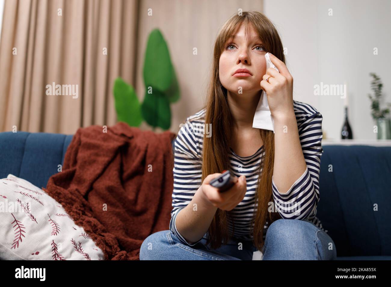 Traurige junge Frau, die mit Seidenpapier und Fernbedienung in der Hand auf dem Sofa sitzt und einen Kinofilm im Fernsehen sieht Stockfoto
