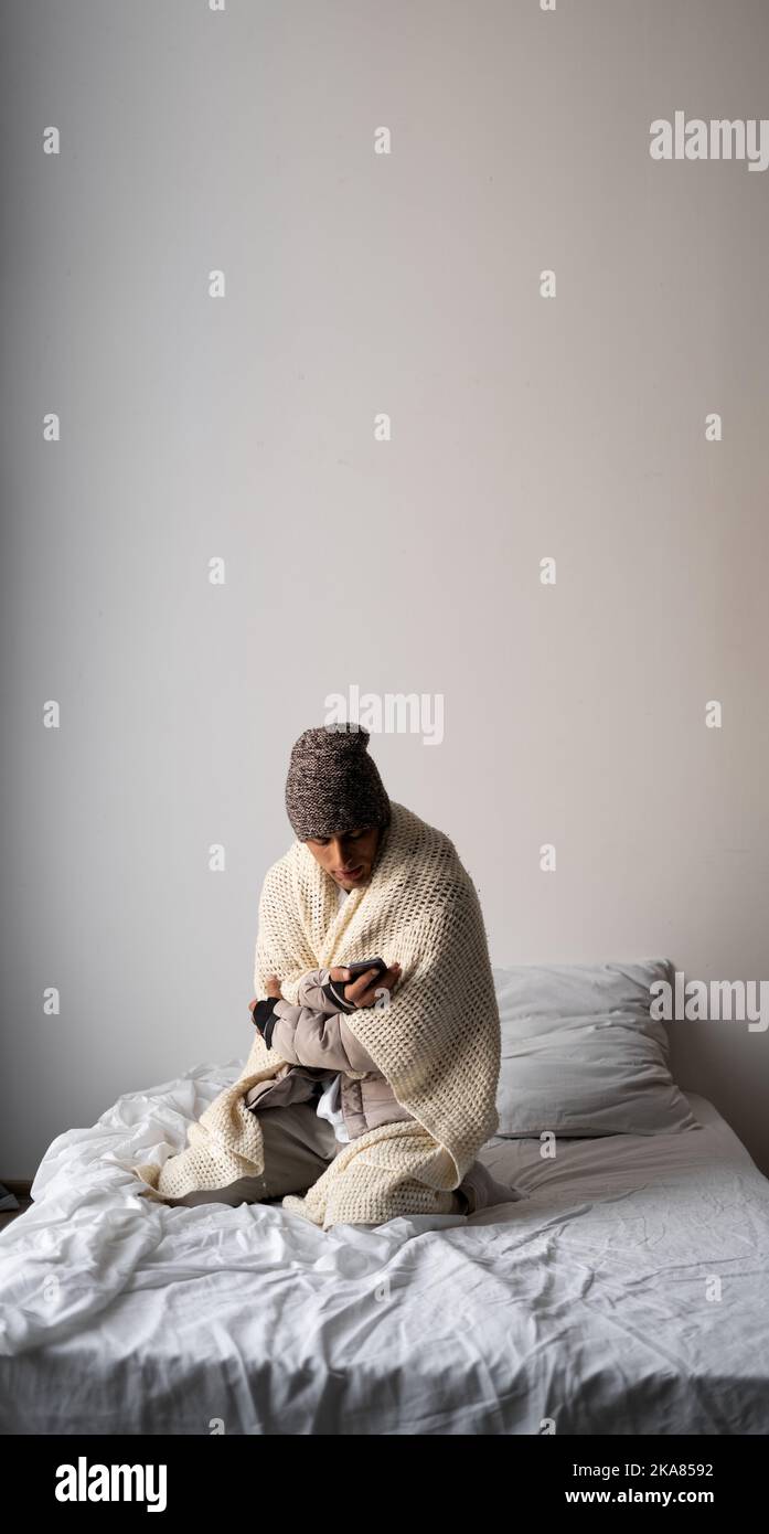 Der junge Mann, der warm angezogen ist, fühlt sich kalt an, bedeckt mit warmen Karli, sitzt auf dem Bett im kalten Zuhause. Haben Sie Probleme mit der Gesundheit, Zentralheizung Stockfoto