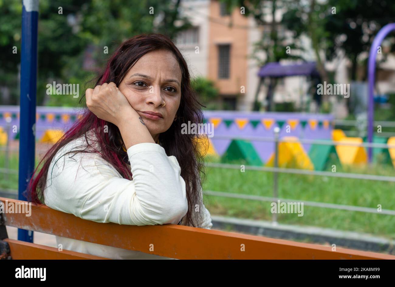 Mid Adult Sorgen Frau sitzt auf der Hand auf dem Kinn Pose in einer Parkbank und Blick auf eine Entfernung. Vorderansicht. Indische Ethnizität Alter 50 bis 54 Jahre. Stockfoto