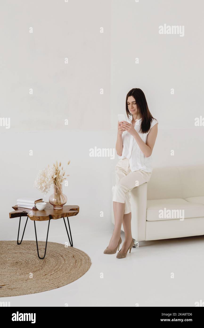 Attraktive Frau in Weiß mit Smartphone, während sie auf dem Rand des Sofas sitzt. Stockfoto
