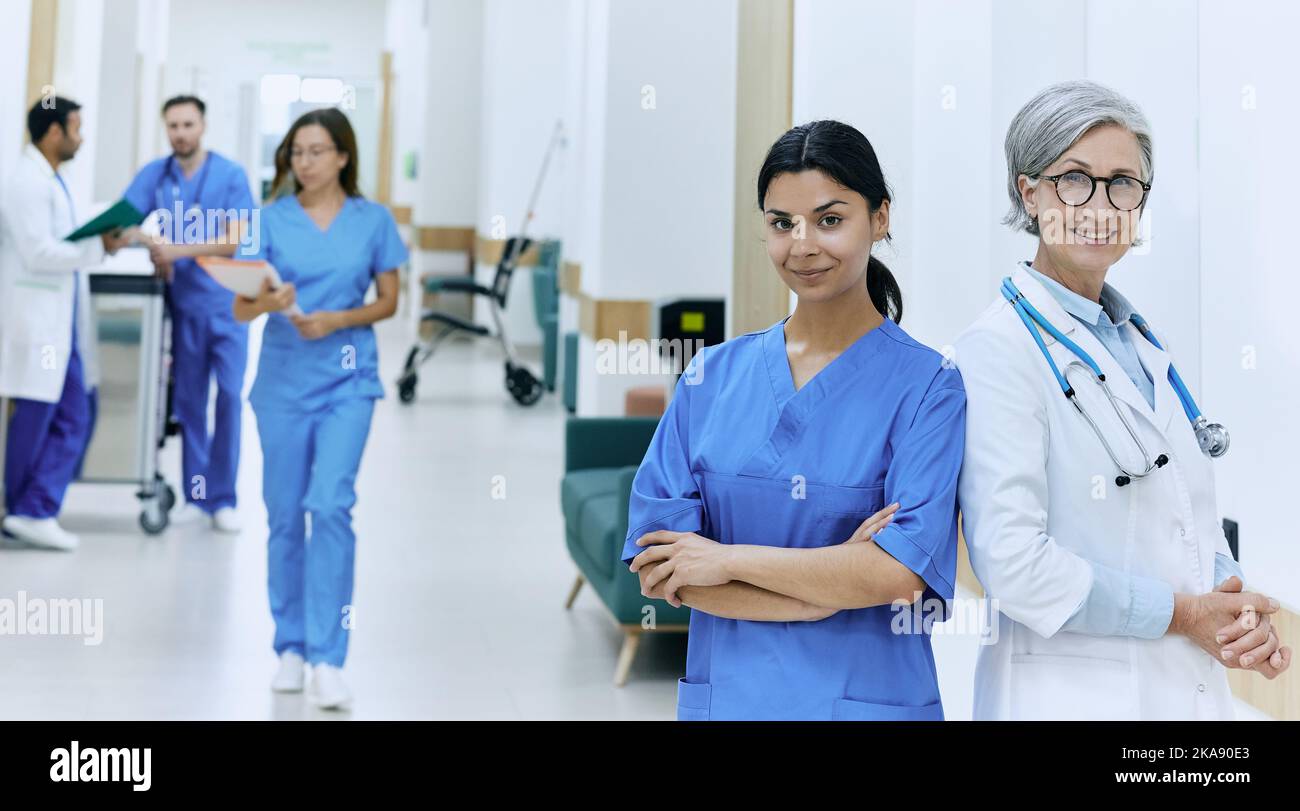 Erfahrener Chefarzt posiert mit asiatischer Krankenschwester vor dem Hintergrund des medizinischen Personals, der den Korridor der medizinischen Klinik entlang läuft. Medizinisches Team Stockfoto
