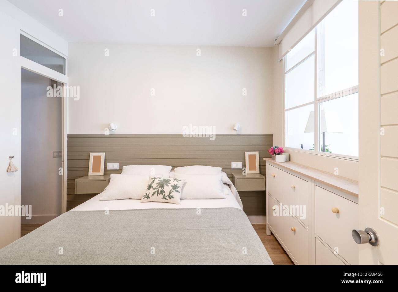 Schönes, aber enges Schlafzimmer mit einer weißen Kommode, grauem Kopfteil und einem Erkerfenster an einer Seitenwand Stockfoto
