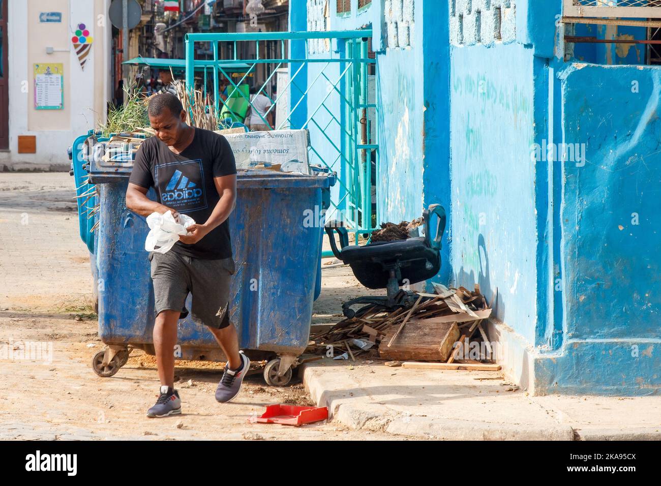 Ein Kubaner läuft an Müllcontainern der Stadt vorbei. Er trägt eine Plastiktüte und trägt ein Adidas T-Shirt Stockfoto