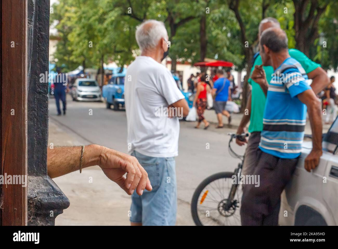 Die Hand eines Mannes, der eine Kette trägt, lehnt sich an eine Säule. Drei weitere Männer sprechen mit einem LADA-Auto. Stockfoto