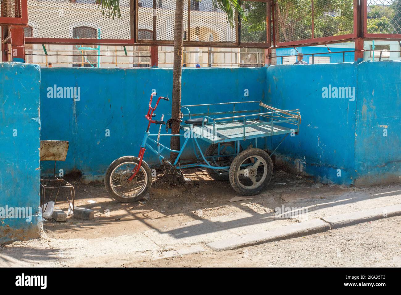 Ein Dreirad ist an einen Metallstab gekettet. Am Zaun des Gebäudes befindet sich auch ein kaputter Stuhl. Stockfoto